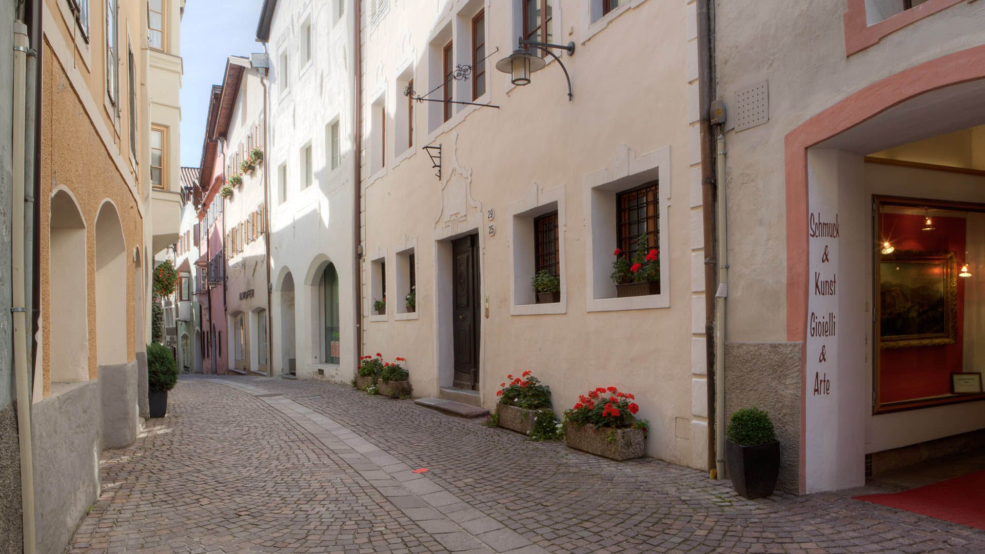 Stradina storica nel centro di Chiusa con edifici colorati, insegne d’arte e fiori ai balconi