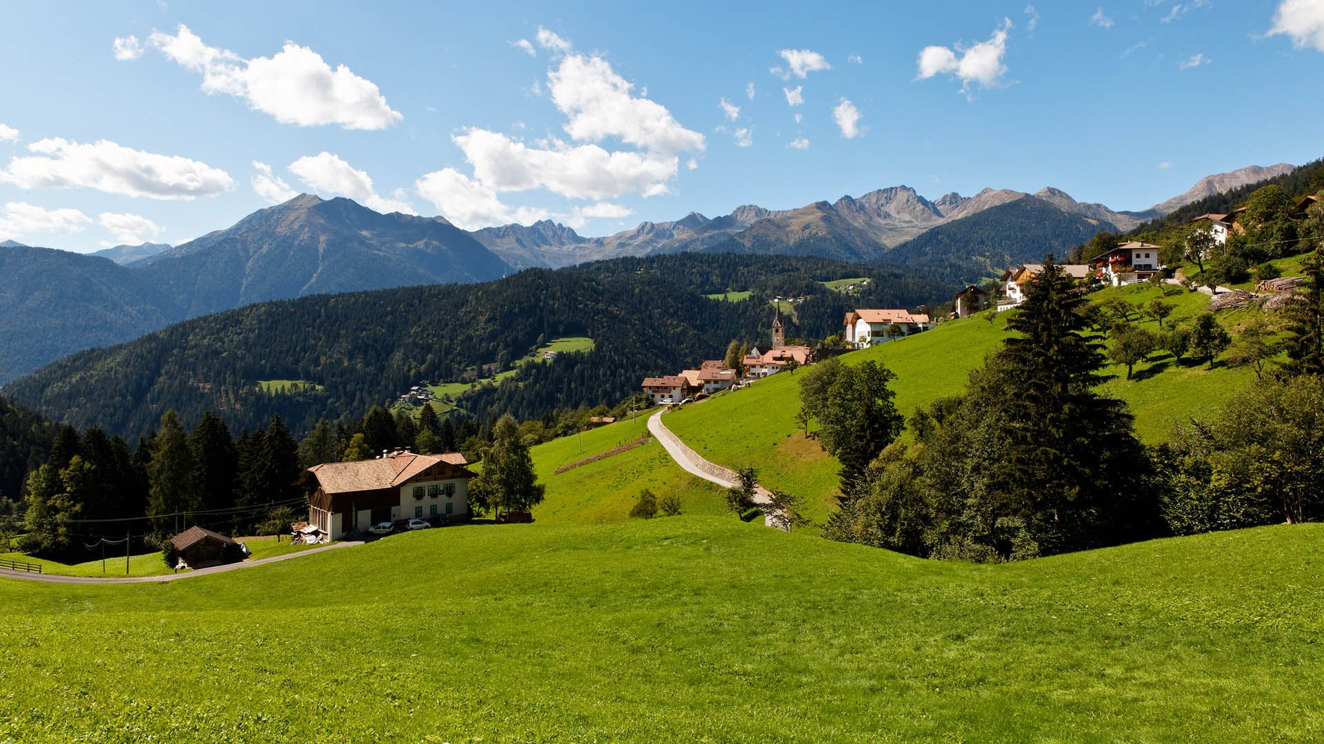 Panorama su Lauregno tra prati verdi e montagne, con chiesa e case alpine sotto il cielo azzurro